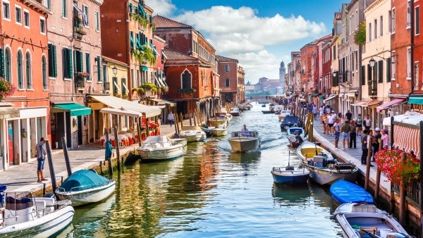 A boat travels along the canals of Venice. The canal is bordered by picturesque buildings and parked boats.