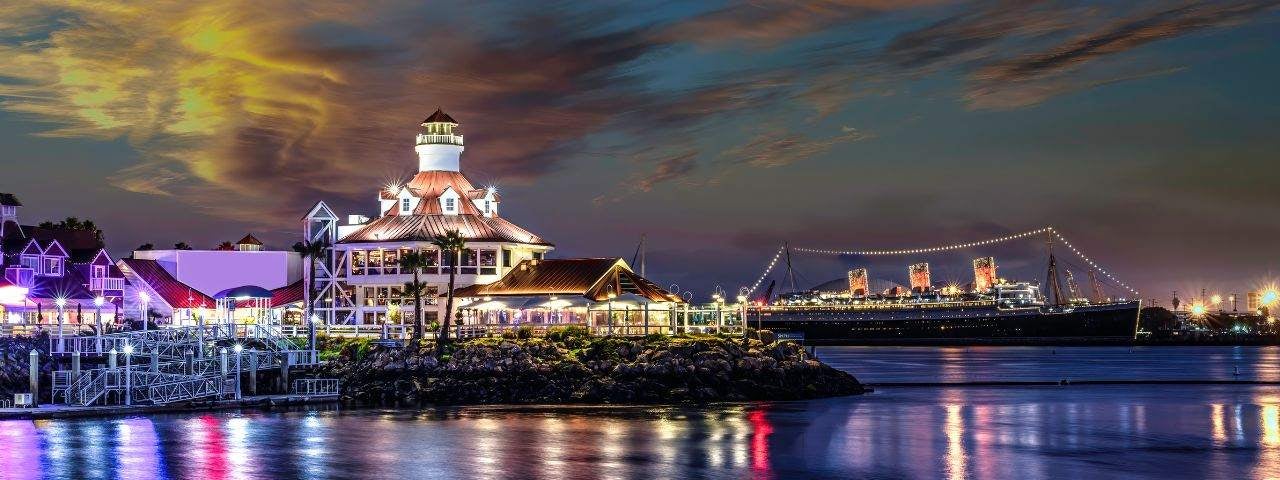 A photograph of the Long Beach Lighthouse near Los Angeles, lit by colorful lights that stand out against the night sky and are reflecting off the ocean. 