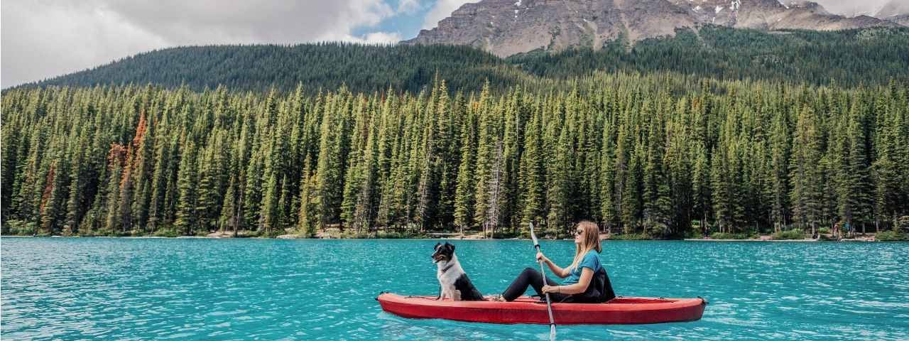A photograph of a blonde girl and her Border Collie dog on a kayak on Moaine Lake in Banff National Park, Canada. 