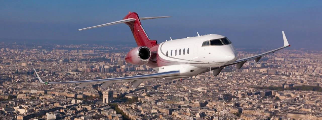 A white and dark red private jet flies high through a clear blue sky over the sprawling cityscape of Paris, with the iconic Arc de Triomphe visible below.