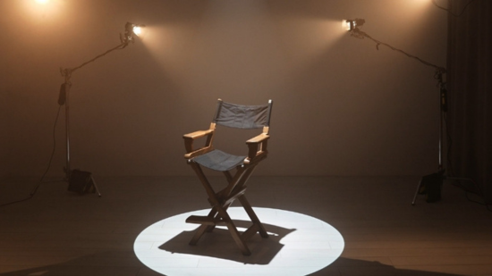 An empty black wooden director's chair positioned in the centre of a stage spotlight on a light wood plank floor, flanked by two vintage studio lights on tall metal stands with dark curtains draping the background in a dimly lit production set. 